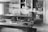 A vintage workshop with wooden paneling features a large workbench covered with various tools and materials. Shelves on the walls hold numerous boxes and supplies, while a hanging lamp provides lighting. The scene conveys a sense of organized clutter typical of a busy workspace.