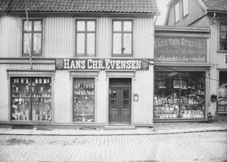 A black and white photograph of an old storefront with the name Hans Chr. Evensen displayed prominently. The building has large windows filled with an assortment of goods, including hardware and household items. The structure appears to be made of wood, with a tiled roof and cobblestone street in front.