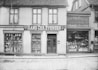 A black and white photograph of an old storefront with the name Hans Chr. Evensen displayed prominently. The building has large windows filled with an assortment of goods, including hardware and household items. The structure appears to be made of wood, with a tiled roof and cobblestone street in front.