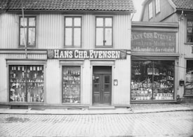 A black and white photograph of an old storefront with the name Hans Chr. Evensen displayed prominently. The building has large windows filled with an assortment of goods, including hardware and household items. The structure appears to be made of wood, with a tiled roof and cobblestone street in front.