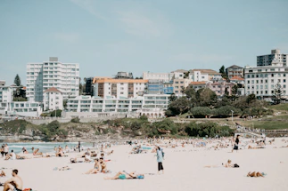 A crowded beach scene features numerous people relaxing on the sand. In the background, a row of modern and older-style apartment buildings is visible, along with some greenery and trees. The sky is clear, adding to the sunny and warm atmosphere.