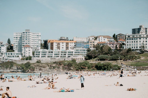 A crowded beach scene features numerous people relaxing on the sand. In the background, a row of modern and older-style apartment buildings is visible, along with some greenery and trees. The sky is clear, adding to the sunny and warm atmosphere.