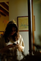 A person wearing a golden tiara stands near a window, looking down at a smartphone in their hands. The room is dimly lit, with soft light entering from the window. The walls have some framed artwork, and the ceiling features wooden beams.