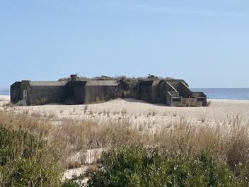 A concrete bunker situated on a sandy beach, partially covered with overgrown grass and vegetation. The ocean is visible in the background with a clear blue sky above, and the surrounding area has sparse dune grass.