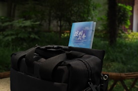 A black bag is placed on a stone surface, with a blue pamphlet or brochure partially visible from one of its pockets. The background reveals a lush, green outdoor setting with foliage and trees, suggesting a park or garden area.