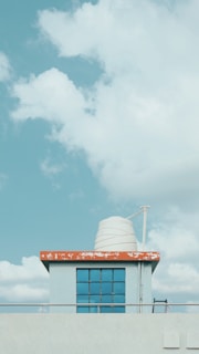 A small rooftop structure with a red and white edge roof and a white water tank on top. The building has a window with a blue tint and is set against a backdrop of a blue sky filled with fluffy white clouds.