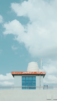 A small rooftop structure with a red and white edge roof and a white water tank on top. The building has a window with a blue tint and is set against a backdrop of a blue sky filled with fluffy white clouds.
