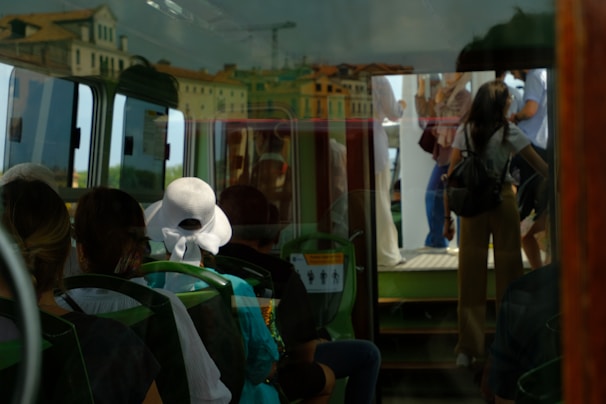 Passengers are seated inside a vehicle with green seats visible. The reflection on the window shows people standing outside, along with the facade of nearby buildings featuring classic architecture. A woman wearing a white hat sits among the passengers, adding a touch of elegance to the scene.