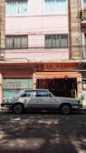 A street scene with a classic white sedan parked in front of a storefront with a sign reading 'La Argentina'. The building has a pastel pink facade with windows featuring security bars and curtains. The shop displays various items and is flanked by closed, graffiti-covered metal shutters.