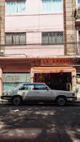 A street scene with a classic white sedan parked in front of a storefront with a sign reading 'La Argentina'. The building has a pastel pink facade with windows featuring security bars and curtains. The shop displays various items and is flanked by closed, graffiti-covered metal shutters.