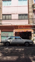 A street scene with a classic white sedan parked in front of a storefront with a sign reading 'La Argentina'. The building has a pastel pink facade with windows featuring security bars and curtains. The shop displays various items and is flanked by closed, graffiti-covered metal shutters.