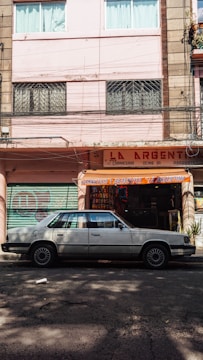 A street scene with a classic white sedan parked in front of a storefront with a sign reading 'La Argentina'. The building has a pastel pink facade with windows featuring security bars and curtains. The shop displays various items and is flanked by closed, graffiti-covered metal shutters.