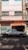 A street scene with a classic white sedan parked in front of a storefront with a sign reading 'La Argentina'. The building has a pastel pink facade with windows featuring security bars and curtains. The shop displays various items and is flanked by closed, graffiti-covered metal shutters.
