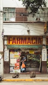 A street view of a pharmacy with a large sign reading 'Farmacia' above the entrance. The shop front is decorated with various posters and advertisements. A man carrying an orange bag is entering the shop. The facade of the building is brown and white with visible electrical wires and a sporadic display of greenery.