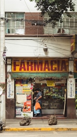 A street view of a pharmacy with a large sign reading 'Farmacia' above the entrance. The shop front is decorated with various posters and advertisements. A man carrying an orange bag is entering the shop. The facade of the building is brown and white with visible electrical wires and a sporadic display of greenery.