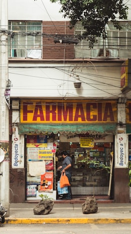A street view of a pharmacy with a large sign reading 'Farmacia' above the entrance. The shop front is decorated with various posters and advertisements. A man carrying an orange bag is entering the shop. The facade of the building is brown and white with visible electrical wires and a sporadic display of greenery.