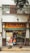 A street view of a pharmacy with a large sign reading 'Farmacia' above the entrance. The shop front is decorated with various posters and advertisements. A man carrying an orange bag is entering the shop. The facade of the building is brown and white with visible electrical wires and a sporadic display of greenery.