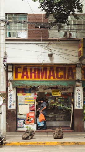 A street view of a pharmacy with a large sign reading 'Farmacia' above the entrance. The shop front is decorated with various posters and advertisements. A man carrying an orange bag is entering the shop. The facade of the building is brown and white with visible electrical wires and a sporadic display of greenery.