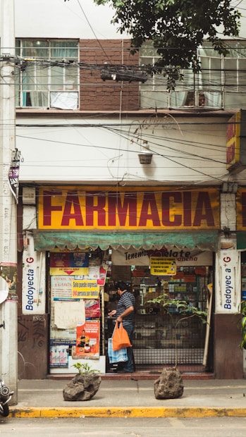A street view of a pharmacy with a large sign reading 'Farmacia' above the entrance. The shop front is decorated with various posters and advertisements. A man carrying an orange bag is entering the shop. The facade of the building is brown and white with visible electrical wires and a sporadic display of greenery.