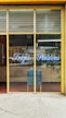 The image depicts a storefront with large glass windows and a yellow frame. On the glass, there is blue script text that reads 'Trajes Pantalones'. Inside, there is a blue chair against a wooden cabinet. A potted plant is visible to the right, and various items can be seen reflected in the glass, including some greenery outside.