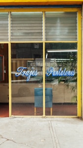 The image depicts a storefront with large glass windows and a yellow frame. On the glass, there is blue script text that reads 'Trajes Pantalones'. Inside, there is a blue chair against a wooden cabinet. A potted plant is visible to the right, and various items can be seen reflected in the glass, including some greenery outside.