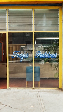 The image depicts a storefront with large glass windows and a yellow frame. On the glass, there is blue script text that reads 'Trajes Pantalones'. Inside, there is a blue chair against a wooden cabinet. A potted plant is visible to the right, and various items can be seen reflected in the glass, including some greenery outside.