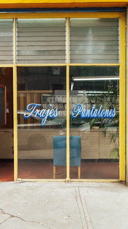 The image depicts a storefront with large glass windows and a yellow frame. On the glass, there is blue script text that reads 'Trajes Pantalones'. Inside, there is a blue chair against a wooden cabinet. A potted plant is visible to the right, and various items can be seen reflected in the glass, including some greenery outside.