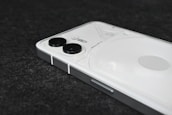A modern smartphone with a sleek design, featuring a white back panel and dual cameras positioned vertically in the top left corner. The device is placed on a dark textured surface.