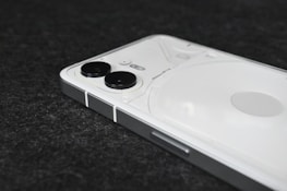 A modern smartphone with a sleek design, featuring a white back panel and dual cameras positioned vertically in the top left corner. The device is placed on a dark textured surface.