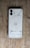 A smartphone placed face down on a wooden surface, revealing a transparent back showcasing internal components like coils and screws. The phone has a modern, minimalist design with dual cameras and clean lines.
