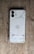 A smartphone placed face down on a wooden surface, revealing a transparent back showcasing internal components like coils and screws. The phone has a modern, minimalist design with dual cameras and clean lines.