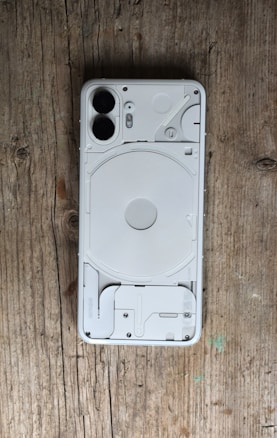 A smartphone placed face down on a wooden surface, revealing a transparent back showcasing internal components like coils and screws. The phone has a modern, minimalist design with dual cameras and clean lines.