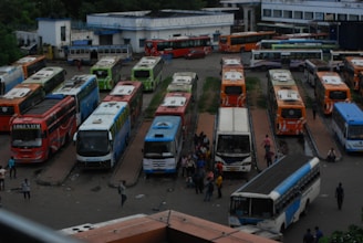 A bustling bus terminal with numerous buses parked in designated bays. The buses are of various colors, including red, blue, green, and orange. People are scattered throughout the terminal, some walking between the buses, while others are gathered in small groups. The terminal buildings are visible in the background, adding to the urban setting.