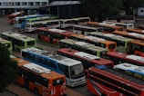 A large parking area filled with numerous colorful buses, arranged in neat rows. The buses are of various colors including orange, red, blue, and green. The scene appears to be at a bus depot or terminal, surrounded by some trees and buildings in the background.