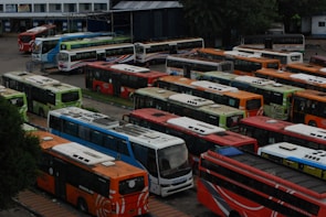 A large parking area filled with numerous colorful buses, arranged in neat rows. The buses are of various colors including orange, red, blue, and green. The scene appears to be at a bus depot or terminal, surrounded by some trees and buildings in the background.