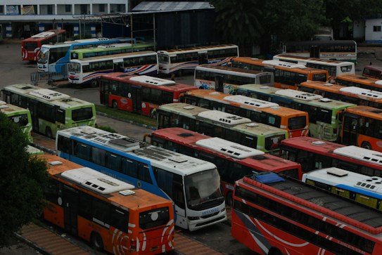 A large parking area filled with numerous colorful buses, arranged in neat rows. The buses are of various colors including orange, red, blue, and green. The scene appears to be at a bus depot or terminal, surrounded by some trees and buildings in the background.