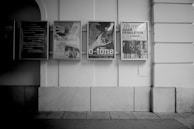 A row of framed posters mounted on a building wall, featuring text and abstract imagery. The scene is in black and white, adding a classic and dramatic touch.