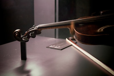 A close-up view of a violin positioned on a dark, reflective surface. The focus is on the scroll and neck of the violin, with the bow placed beside it. Soft lighting casts shadows, enhancing the rich brown tones of the wood.
