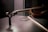 A close-up view of a violin positioned on a dark, reflective surface. The focus is on the scroll and neck of the violin, with the bow placed beside it. Soft lighting casts shadows, enhancing the rich brown tones of the wood.