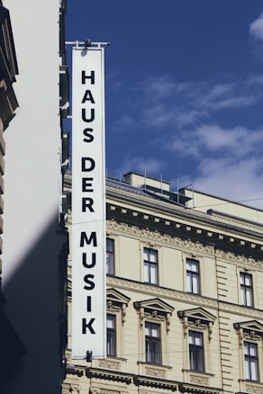 A tall building with ornate architectural details and a large vertical sign that reads 'Haus der Musik' hangs from its facade. The sky is blue with a few clouds, creating a contrast with the cream-colored building.