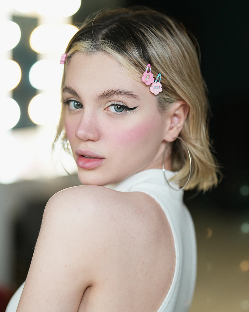 Margaux in a white halter top at a dressing-room mirror ringed with bulbs, looking back over her shoulder.