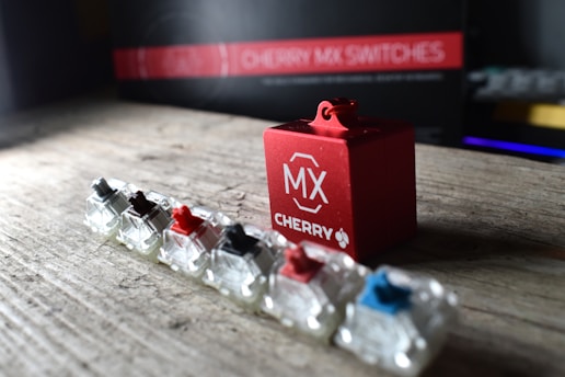 A set of mechanical keyboard switches, each with different colored stems, is lined up on a wooden surface. A prominent red cube labeled 'MX CHERRY' is positioned beside them, and in the background, there is a blurred image of a box with text indicating 'CHERRY MX SWITCHES.' The lighting highlights the glossy and transparent nature of the switches.