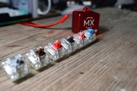 Several mechanical keyboard switches of different colors are arranged on a wooden surface. A small red box with a logo and carrying strap is positioned in the background, slightly blurred. The overall setup appears casual and focused on showcasing the variety of switches.