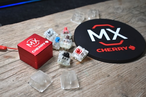 On a wooden surface, multiple mechanical keyboard switches and keycaps are arranged around a small red box with the MX Cherry logo. Nearby, a round black object also displays the MX Cherry branding.