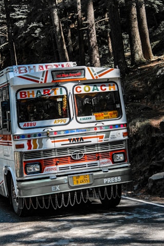 A colorful bus labeled 'Bharti Coach' navigates a winding road through a dense forest. The bus features vibrant decorations and patterns, with its front displaying various inscriptions and logos. Tall trees with thick trunks and lush foliage surround the road, casting shadows on the pavement.