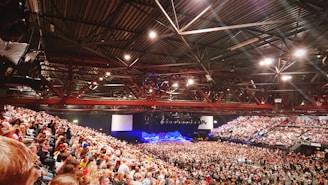 A large indoor concert arena filled with a vast audience. The stage is set up with lights and equipment, and people are sitting in tiered seating as well as standing in front of the stage. The ceiling displays metal beams and lighting fixtures that illuminate the space.