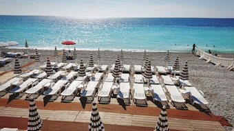 A beach scene with rows of white sun loungers and black-and-white striped umbrellas positioned on a wooden deck in front of a pebble beach. The vibrant blue ocean stretches into the horizon under a clear sky. A few people are swimming in the water and there is one red umbrella among the black-and-white ones.