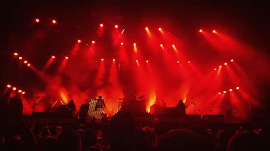 A lively concert scene with intense red stage lighting illuminating musicians and their instruments. The silhouettes of performers are visible against a backdrop of vibrant red haze created by spotlights and smoke effects. The audience can be seen as dark figures at the bottom of the image.