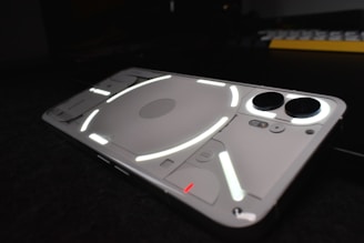 A sleek, modern smartphone with a transparent back showing luminous white and red lines, creating a geometric pattern. The rear camera and other components are visible through the casing, lying on a dark surface with subtle reflections.