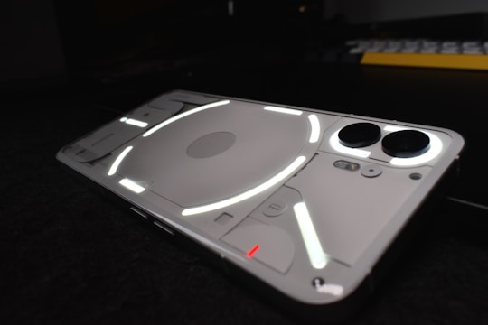 A sleek, modern smartphone with a transparent back showing luminous white and red lines, creating a geometric pattern. The rear camera and other components are visible through the casing, lying on a dark surface with subtle reflections.